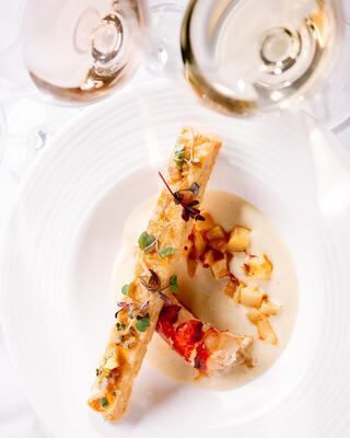 A gourmet dish features lobster in a creamy sauce, paired with golden sautéed apples, garnished with fresh herbs and accompanied by wine glasses.