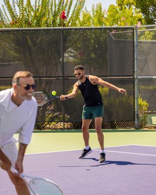 Two men play tennis on a sunny court, one in a tank top preparing to hit while the other, in white, focuses on the game.