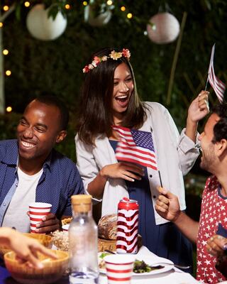 A joyful outdoor gathering features friends celebrating with American flags, festive decorations, and cheerful conversation over food and drinks.