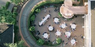 An aerial view of an outdoor gathering shows elegantly set tables surrounded by greenery, with guests mingling and enjoying the atmosphere.