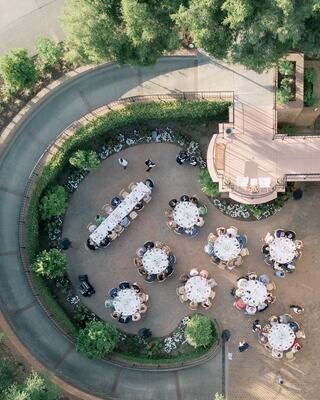 An elegant outdoor gathering features round tables set for dining, surrounded by greenery and pathways, creating a sophisticated atmosphere.