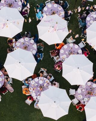 Aerial view of tables adorned with purple linens and white umbrellas, surrounded by guests in colorful attire enjoying an outdoor gathering.