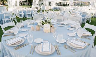 A beautifully set outdoor dining area with light blue tablecloths, elegant tableware, and floral centerpieces for a special event.