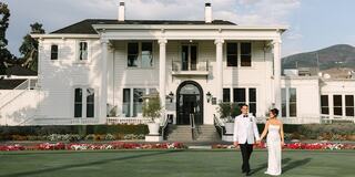 A couple in formal attire strolls hand-in-hand in front of an elegant white mansion, surrounded by vibrant flowers and a scenic landscape.