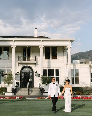 A couple walks hand in hand in formal attire, with a grand white mansion and colorful flowerbeds in the background, under a blue sky.