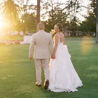 A couple walks hand in hand on a lush green lawn, bathed in warm sunlight, exuding love and joy in their wedding attire.