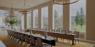 Room with two rows of long tables and chairs set up with a view of trees out the window