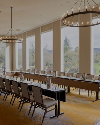 Room with two rows of long tables and chairs set up with a view of trees out the window