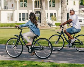 Man and woman riding bikes in front of a mansion on a paved path surrounded by grass