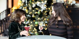 two women clinking glasses in front of a christmas tree
