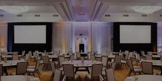 Room set up for a group of people with round tables and chairs, two projector screens, and two chandeliers