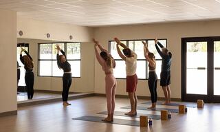 A group of individuals participates in a yoga class, stretching with arms raised, reflected in a large mirror in a bright, airy studio.