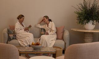 Two women in robes relax on a couch, chatting and enjoying each other's company in a serene, stylish setting with plants and a fruit bowl.