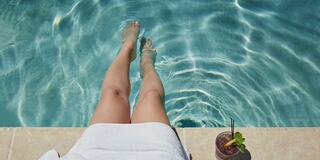 Woman's Legs in Pool While She Sits on the Edge of the Pool Next to a Cocktail
