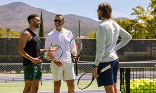 Three men chat on a tennis court, holding rackets, with a sunny backdrop of mountains and trees, creating a relaxed athletic atmosphere.