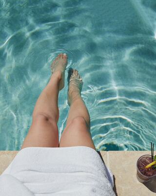 Woman's Legs in Pool While She Sits on the Edge of the Pool Next to a Cocktail