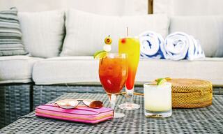 A Variety of Cocktails, a Wallet, and Sunglasses on an Outdoor Patio Table Next to an Outdoor Sofa with Rolled Towels on it
