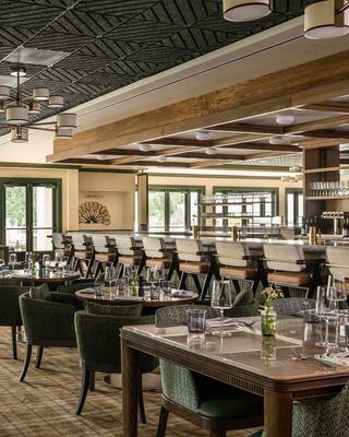 A stylish restaurant features elegant tables, green chairs, and a well-stocked bar, enhanced by warm wood accents and soft lighting.