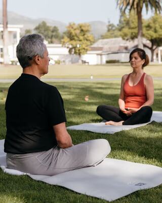 Two practitioners meditate on mats in a serene park, surrounded by greenery and palm trees, embodying calm and focus.