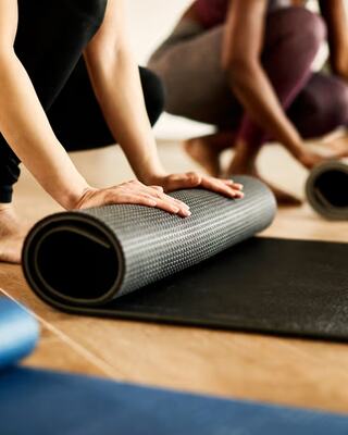 A person is rolling up a yoga mat on a wooden floor, while another person is in a squatting position nearby.