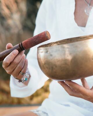 A person holds a wooden mallet above a brass singing bowl, poised to create sound, surrounded by a serene outdoor setting.