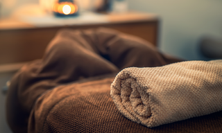 A neatly rolled towel rests on a cozy, textured blanket, with a softly lit background that enhances a tranquil, spa-like atmosphere.