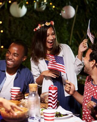 Friends celebrate together, holding small American flags, laughing and enjoying food in a festive outdoor setting with warm lights.