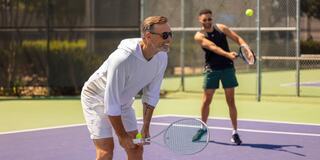Two men play tennis on a sunny court; one prepares to serve while the other readies to receive the ball, both dressed in athletic attire.