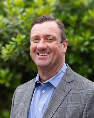 A smiling man in a gray blazer over a blue shirt stands outdoors, surrounded by lush green foliage. His expression is warm and inviting.