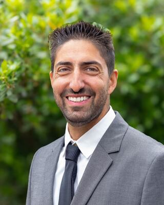 A man in a gray suit with a white shirt and black tie smiles against a backdrop of greenery, exuding confidence and professionalism.