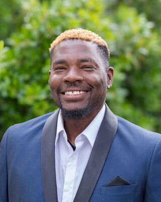 A smiling man in a formal suit with a black lapel and light shirt. He has styled hair with blonde tips, set against lush greenery.
