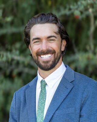 A smiling man with a beard and wavy hair wears a suit jacket and a green tie, posing outdoors against a blurred natural backdrop.