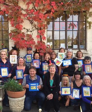 A diverse group of individuals poses with their art creations in front of a building adorned with vibrant autumn leaves.