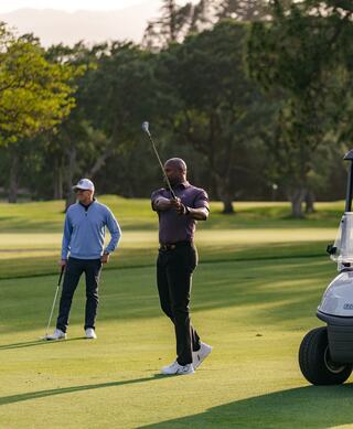 Two golfers on a lush course; one prepares to swing while the other stands nearby, enjoying the serene morning atmosphere.
