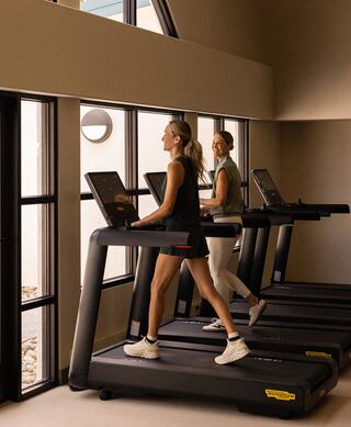 Two women jog on treadmills in a bright, modern gym, smiling and enjoying their workout while sunlight streams through large windows.