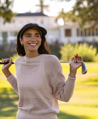 A woman smiles joyfully on a golf course, holding a club and wearing a stylish visor, exuding confidence and a relaxed vibe.