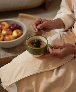 A hand dips a tea bag into a green cup while a bowl of apples sits nearby, creating a cozy, inviting scene.