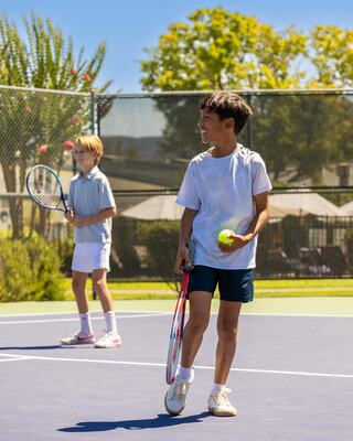 Two boys play tennis on a sunny court, one holding a racket and a ball, while the other prepares to serve. Lush greenery surrounds them.