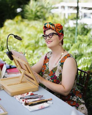 A smiling person with vibrant hair and glasses sits at a garden table, surrounded by art supplies and a sketch pad, enjoying her creative process.