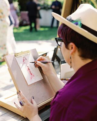An artist, wearing a hat and glasses, sketches a glamorous figure outdoors, surrounded by a lively event atmosphere.
