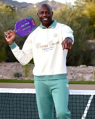 A man poses playfully with a pickleball paddle, wearing a stylish sports outfit, amidst a scenic outdoor setting.