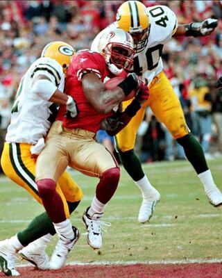 A player in a red jersey makes a catch while being tackled by two opponents in green and yellow uniforms during a football game.