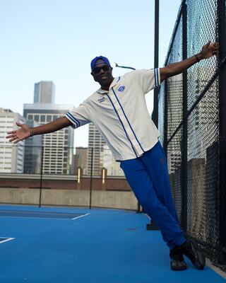 A man in a sporty outfit leans against a fence on a blue court, with city skyscrapers visible in the background.