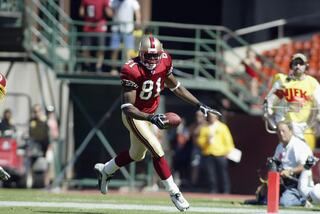 A football player in a red jersey runs with the ball towards the end zone, showcasing skill and determination as fans watch from the stands.