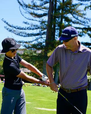 A young golfer receives instruction from an experienced player, with tall trees and a sunny landscape in the background.