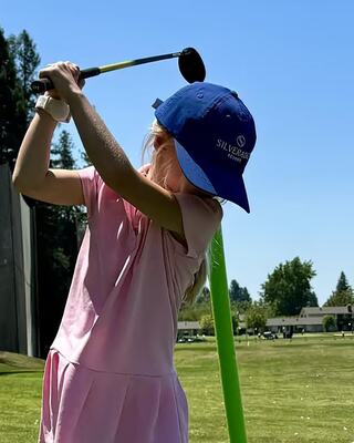 A child in a pink dress and blue cap takes a golf swing at a sunny driving range, surrounded by trees and grassy areas.