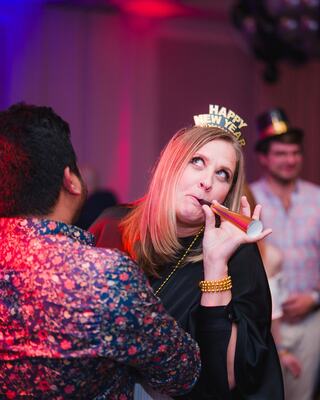 A woman celebrates New Year's Eve, wearing a festive hat and blowing a party horn while dancing with a friend amidst a lively atmosphere.