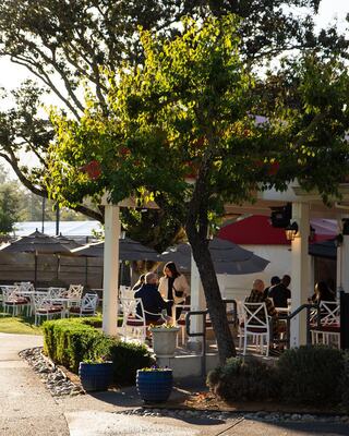 A lively outdoor dining area surrounded by greenery, featuring tables, umbrellas, and guests enjoying the atmosphere in warm sunlight.