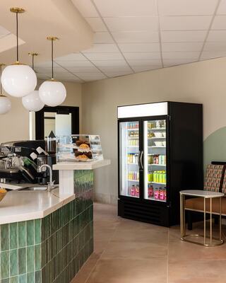 A modern café corner features a coffee machine, a glass display case with pastries, and a refrigerated display filled with beverages.