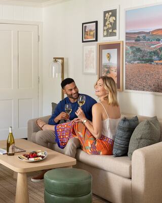 A couple enjoys a cozy moment on a sofa, sharing drinks and smiles, surrounded by warm decor and a view of vineyards.